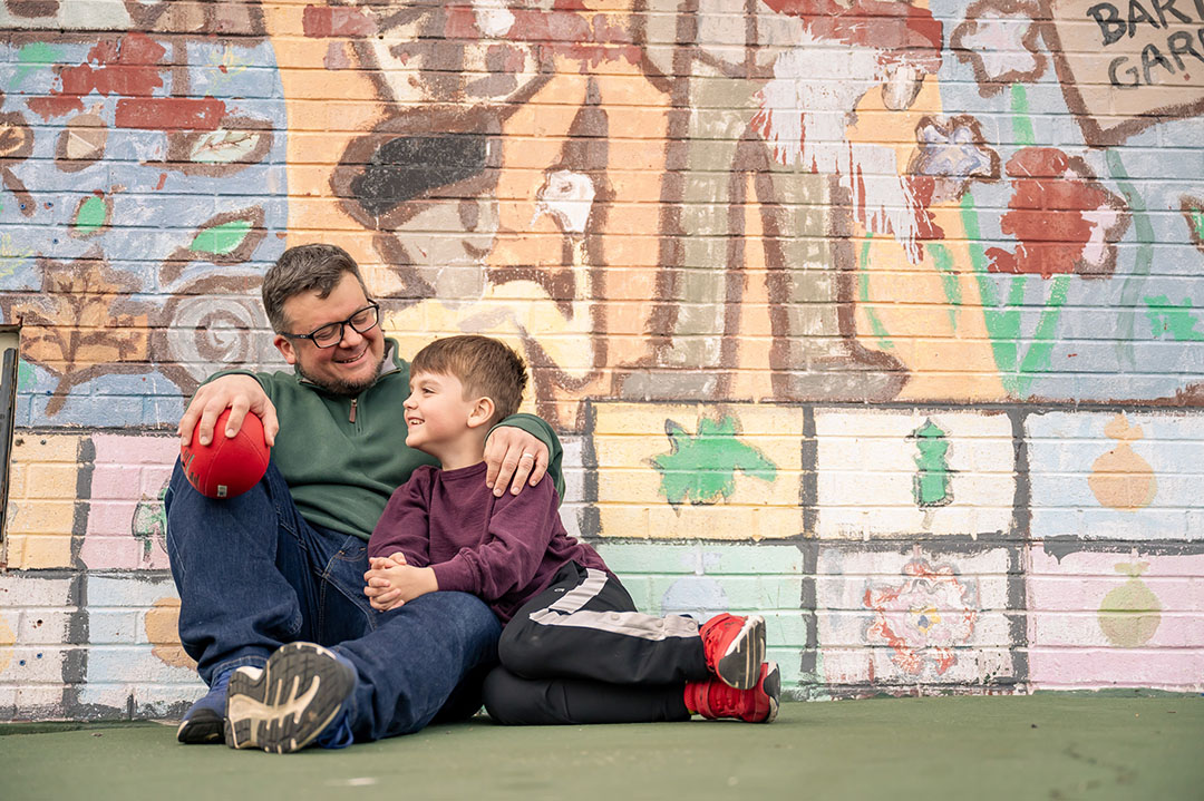 A man and his son sitting together against a wall painted with a colorful mural