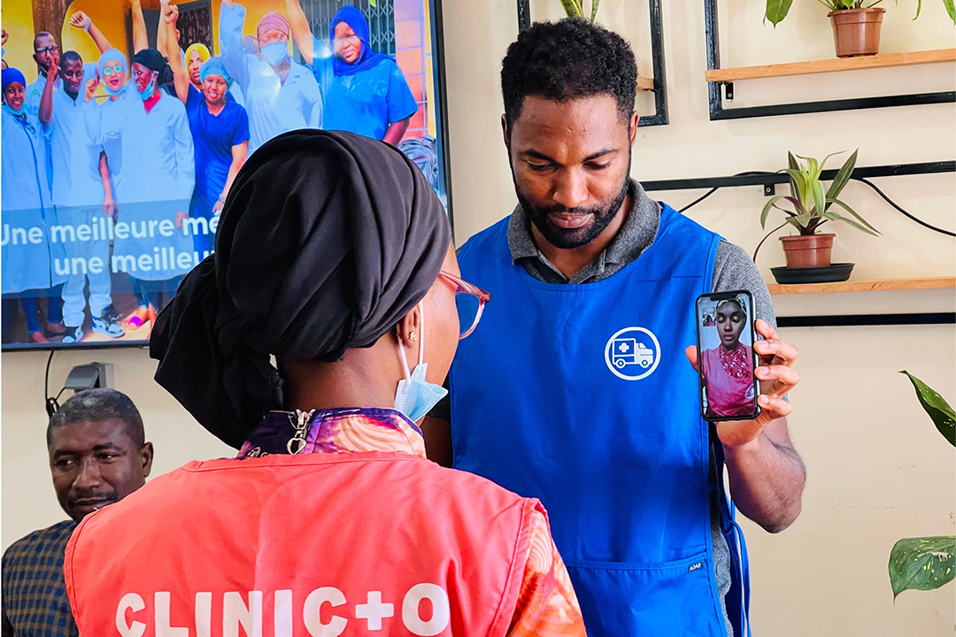 Nasser Diallo in a blue uniform holds up a phone to video conference with a patient