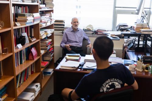 A political science professor at GW in his office sitting across the desk from a student. Shelves of books line the walls.