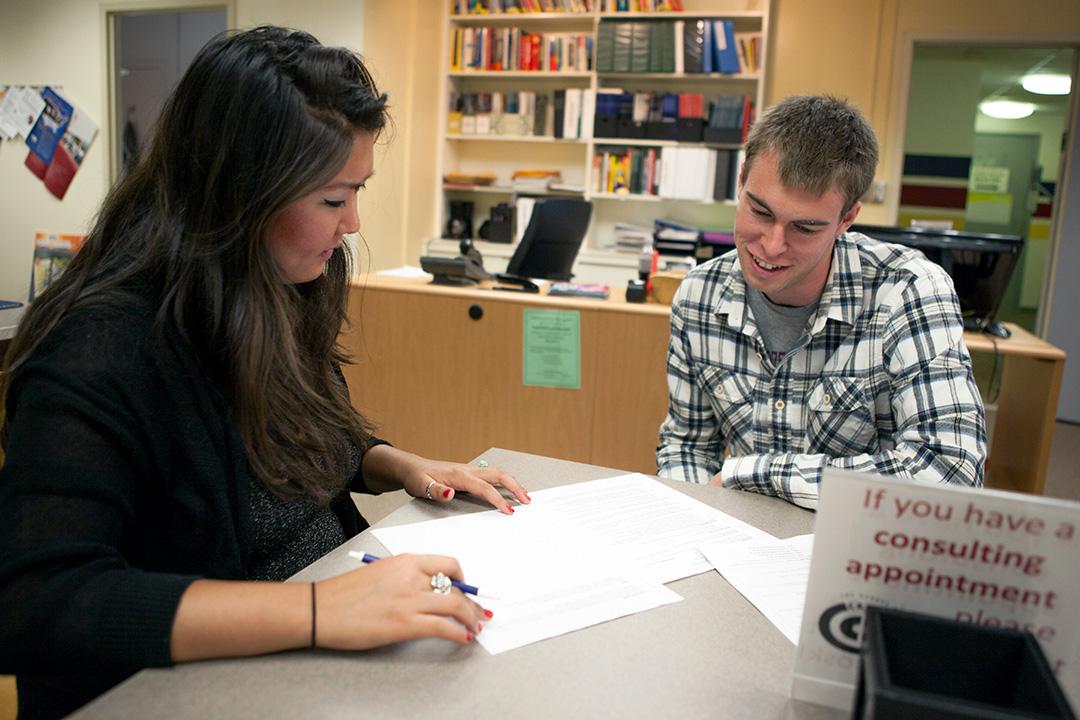 A GW student in a plaid shirt sits at a table with a career counselor looking together at some papers on a table