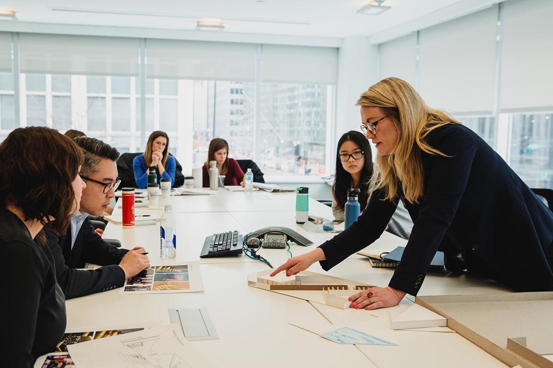 Graduate students in an Interior Architecture classroom lean over a conference table looking at a model, wearing business attire