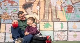 A man and his son sitting together against a wall painted with a colorful mural