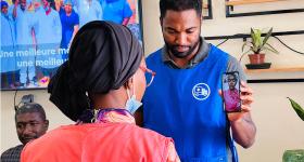 Nasser Diallo in a blue uniform holds up a phone to video conference with a patient